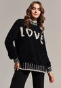 A woman with wavy brown hair wears a black Frogbox by Princess Grobstrickpullover aus Alpaka Wolle Mischung with bold white LOVE lettering and contrast stitching, standing confidently against a plain beige background and facing the camera.