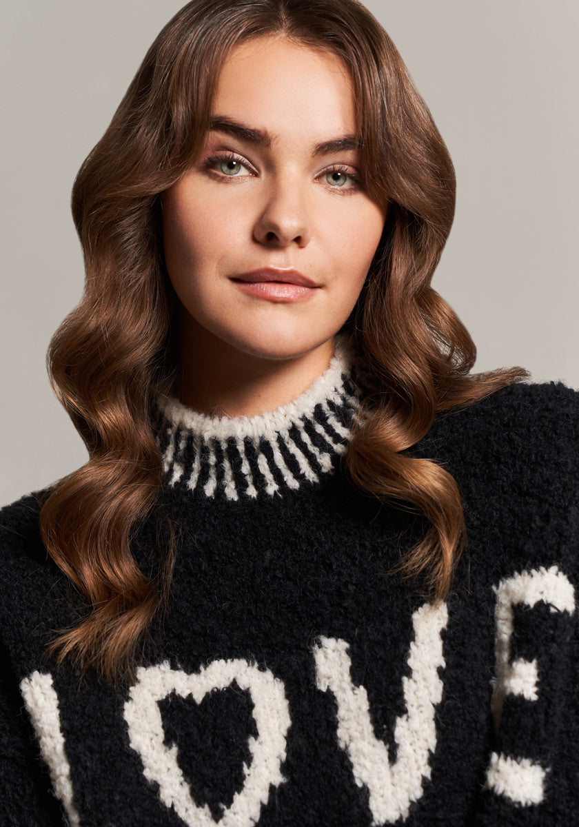 A woman with wavy brown hair and green eyes wears a Frogbox by Princess schwarzer Grobstrickpullover aus Alpaka-Wollmischung mit weißem „LOVE“-Schriftzug, wobei das O als Herz gestaltet ist. Sie blickt sanft in die Kamera, neutral grauer Hintergrund.