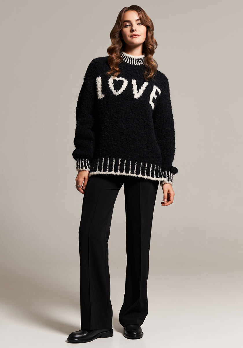 A woman stands in a studio wearing a black Frogbox by Princess Grobstrickpullover with “LOVE” in white letters. Made from an Alpaka Wolle Mischung, the sweater has Kontrastnähte at collar, cuffs, and hem. She pairs it with black trousers and shoes.