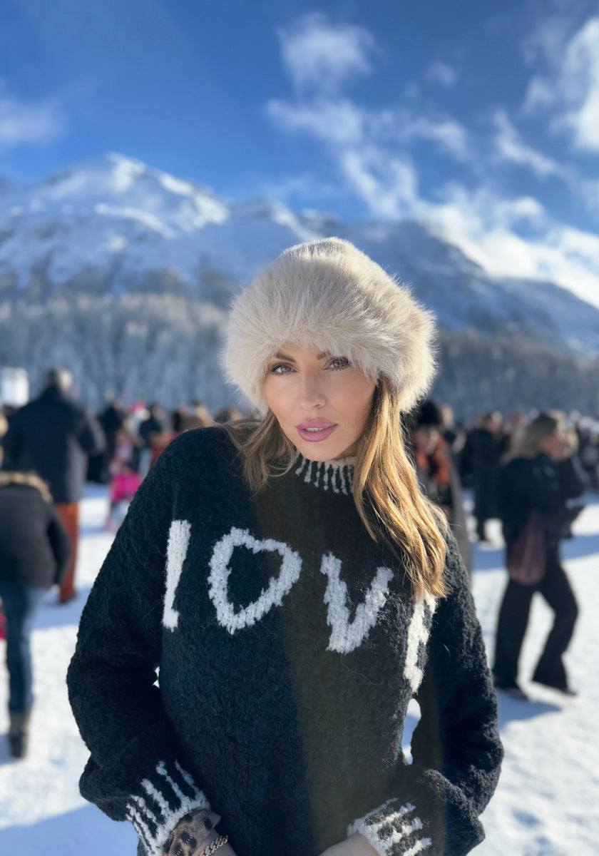 A woman stands in the snow wearing a black Frogbox by Princess Grobstrickpullover with “LOVE” in white letters and a furry beige hat. She poses before a mountain and blue sky as people in winter clothes gather behind her.