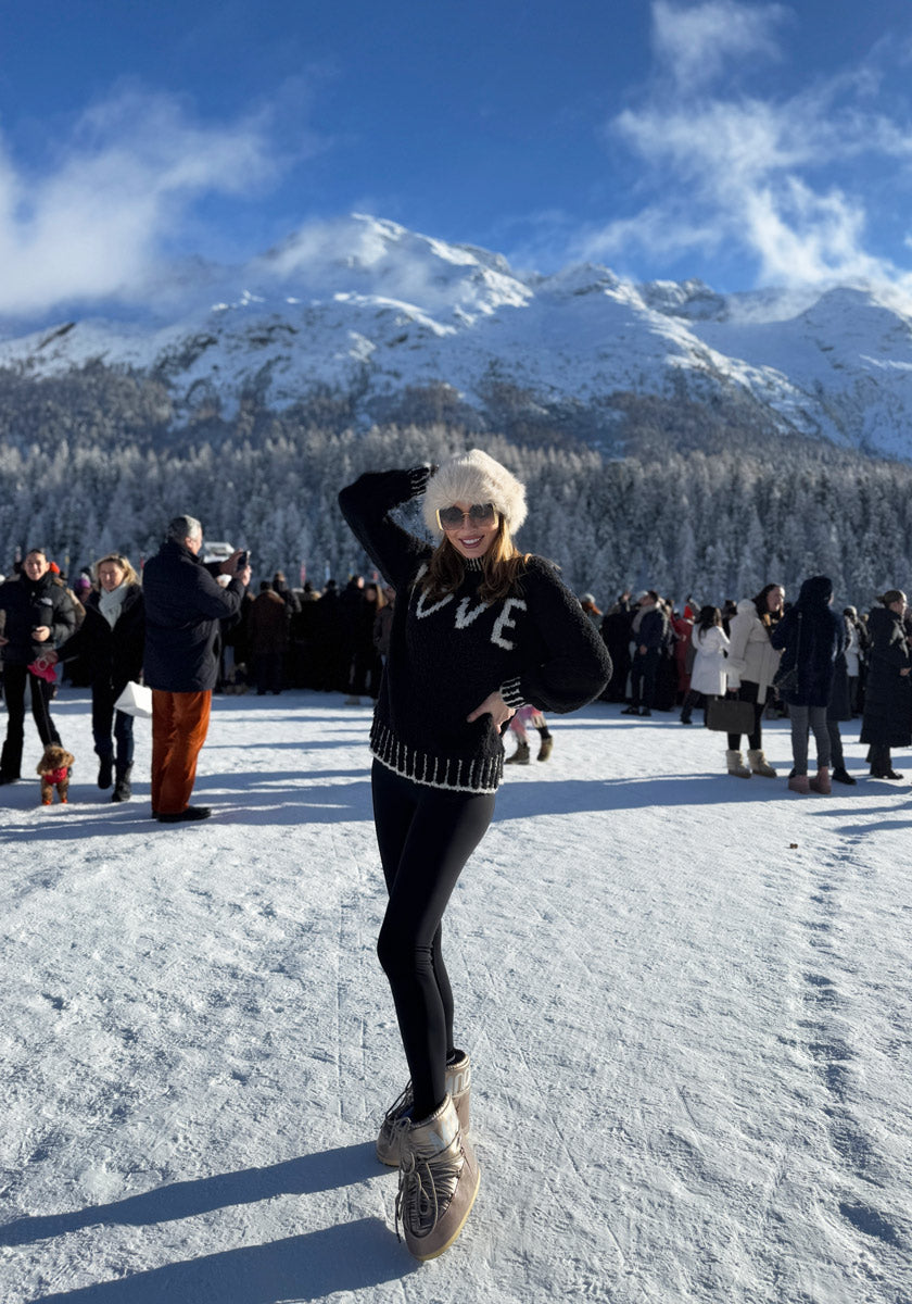 A smiling woman ice skates outdoors on a sunny winter day, wearing a black Frogbox by Princess Grobstrickpullover with Kontrastnähte, white furry hat, sunglasses, and silver skates. Snowy mountains and trees are in the background as others gather nearby.