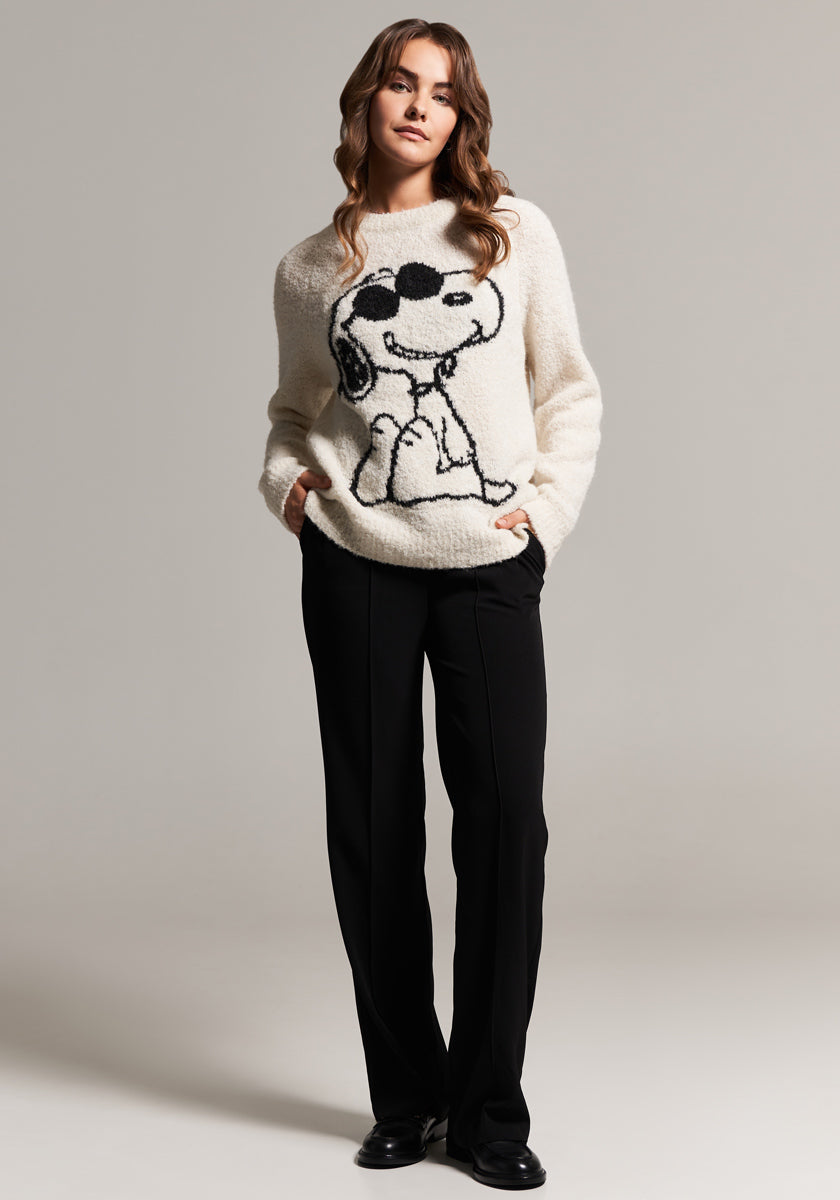 A woman with wavy brown hair wears a cozy Frogbox by Princess Snoopy Pullover made from fuzzy alpaca wool, featuring a bold black Snoopy outline with sunglasses. She pairs it with black straight-leg pants, standing against a light gray background.