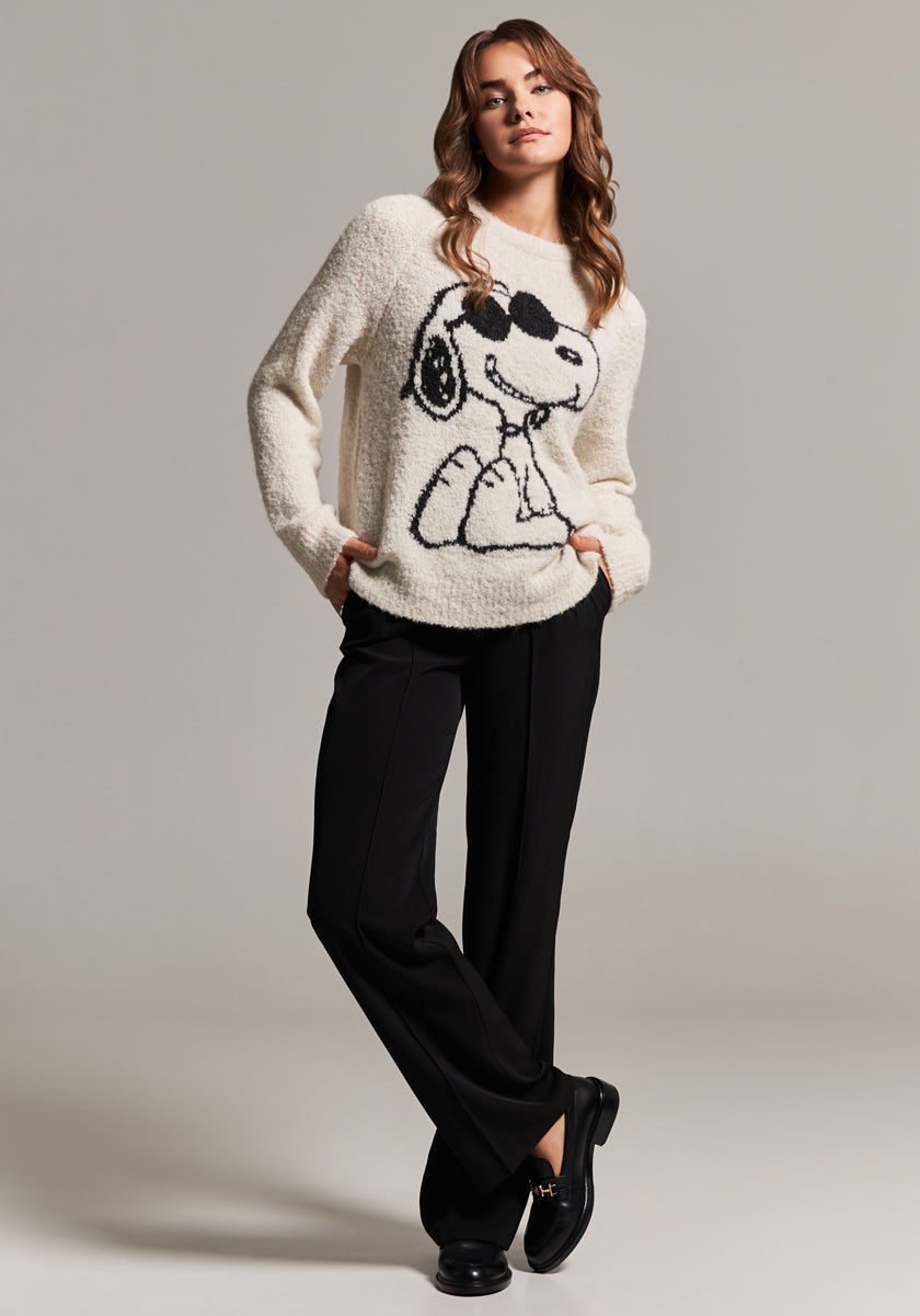 A woman with wavy brown hair poses confidently in a cream-colored Frogbox by Princess Snoopy Grobstrickpullover with a bold black Snoopy outline and sunglasses, black pants, and black loafers against a light gray background.