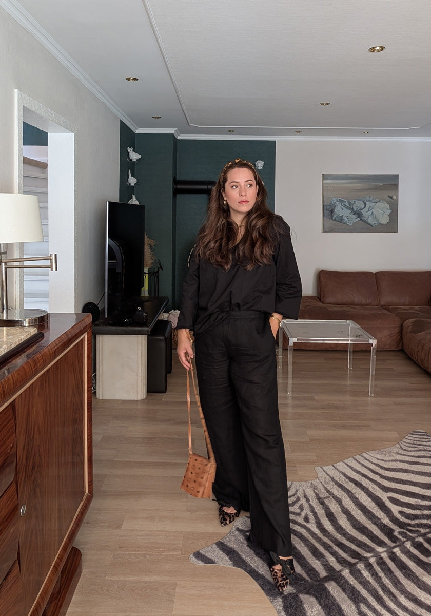 A woman with long brown hair stands in a modern living room, wearing a Frogbox by Princess schwarze Leinen Bluse mit V-Ausschnitt und langen weiten Ärmeln, wide-leg pants, leopard print shoes, and holding a tan crossbody bag.