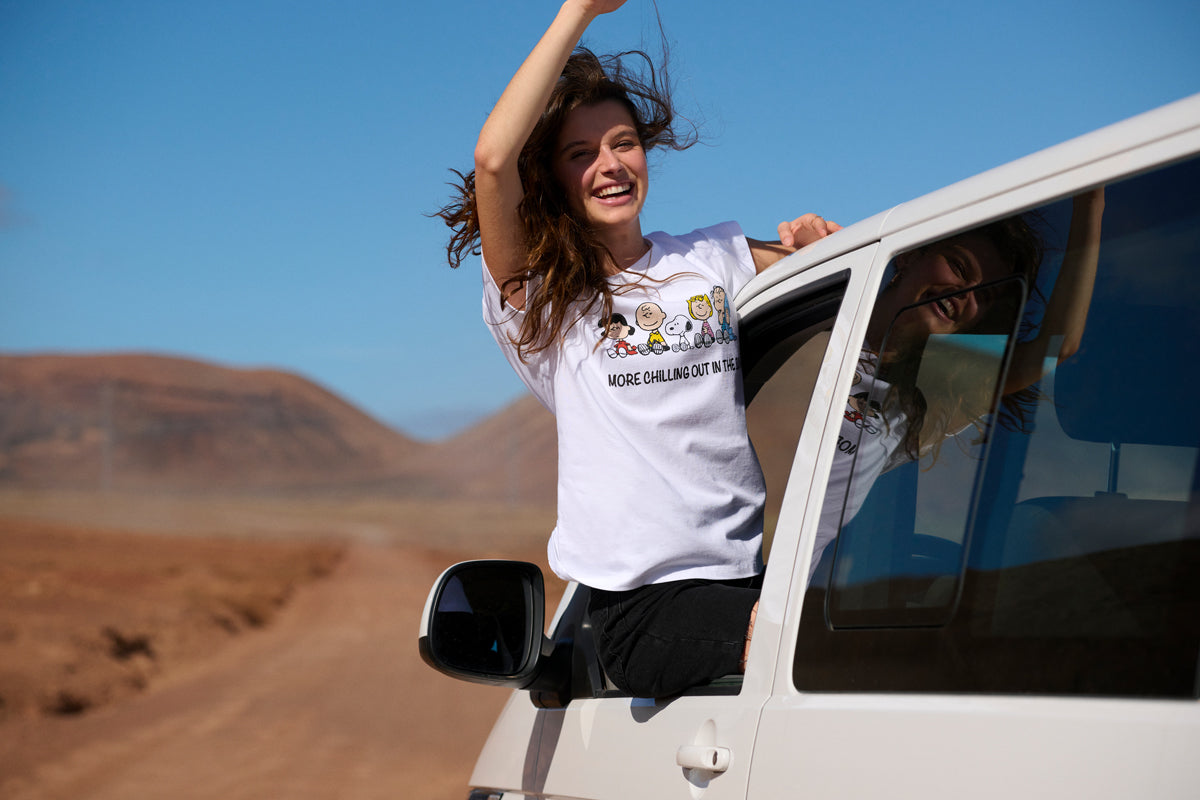 A smiling woman leans out of a white van’s window on a dirt road in a desert landscape. She wears a white T-shirt with cartoon characters and text, raising one arm joyfully. Brown hills and a bright blue sky are visible in the background.