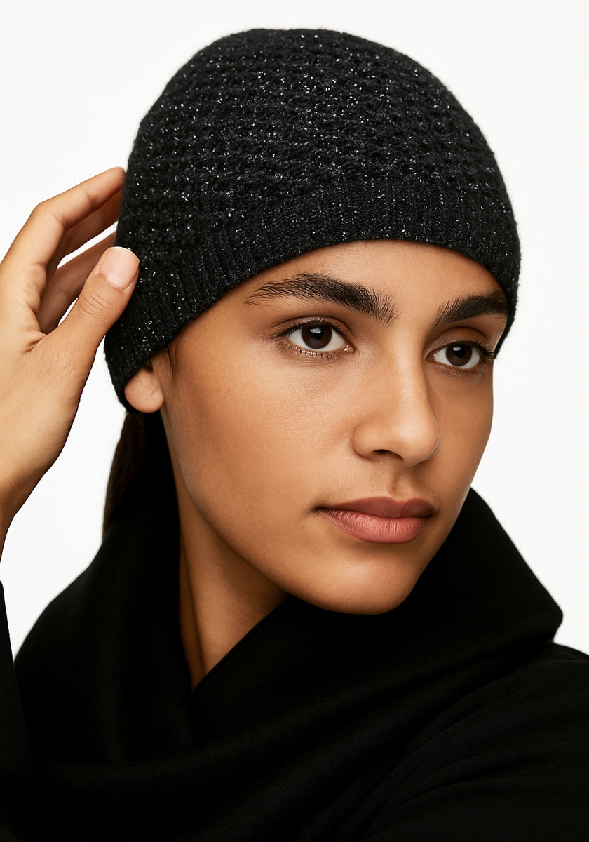 A person with medium skin tone wears a black Princess Goes Hollywood Strickmütze made from a Merino Alpaka blend and matching top. Their dark hair is tucked under the hat as they touch its side, set against a white background, looking calm and neutral.