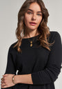 A woman with wavy brown hair wears a black Princess Goes Hollywood Kaschmir Pullover with Rundhals against a plain light background. She accessorizes with layered gold necklaces and a gold bracelet, gazing at the camera with a relaxed, neutral expression.