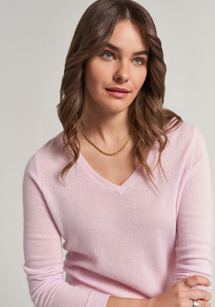 A woman with wavy brown hair and green eyes wears a light pink Princess Goes Hollywood Kaschmir V-Ausschnitt Pullover and a gold chain necklace. She has natural makeup and poses against a plain light background, looking softly to the side.