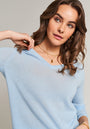 A woman with wavy brown hair wears a light blue Princess Goes Hollywood Kaschmir V-Ausschnitt Pullover, holding her collar while showcasing layered gold necklaces and hoop earrings. She gazes softly to the side against a neutral background.