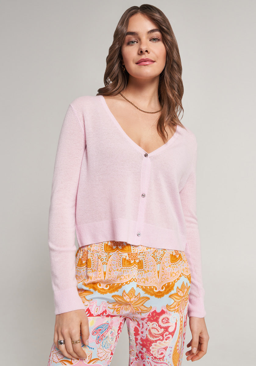 A woman with wavy brown hair wears a light pink V-neck Kaschmir Strickjacke by Princess Goes Hollywood and orange, yellow, and pink paisley pants. She stands against a plain light background, smiling softly with her hands at her sides.