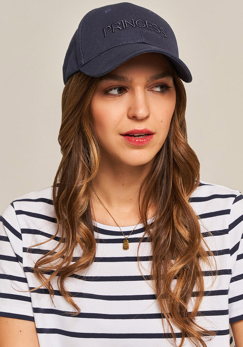 A woman with wavy brown hair wears a blaue Canvas Kappe Princess von Princess Goes Hollywood and a gestreiftes weiß-blaues Baumwoll-T-Shirt. Sie trägt natürliche Schminke, eine goldene Kette und steht vor hellem Hintergrund.