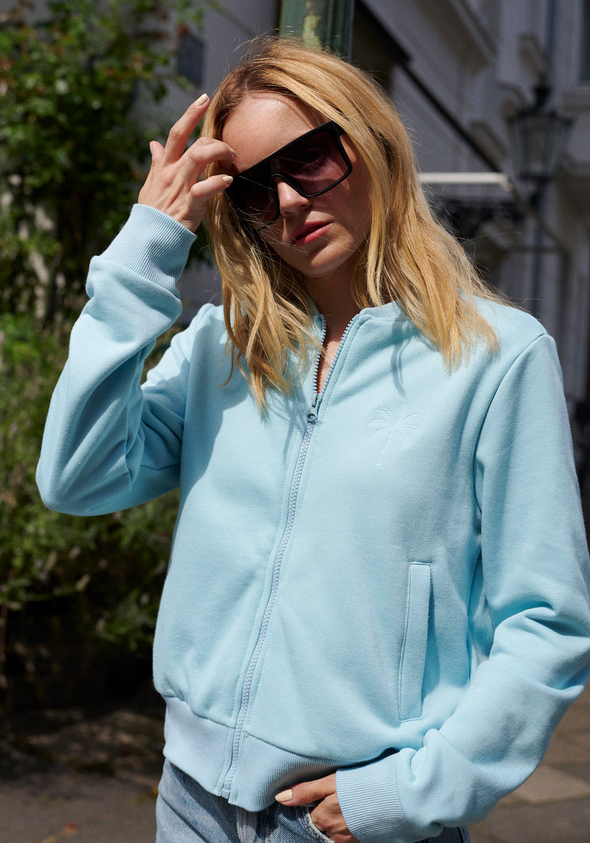 A woman with long blonde hair wears oversized black sunglasses and a light blue Princess Goes Hollywood Sweatjacke with stand-up collar and zipper. She stands outdoors, touching her hair, in front of greenery and building details.