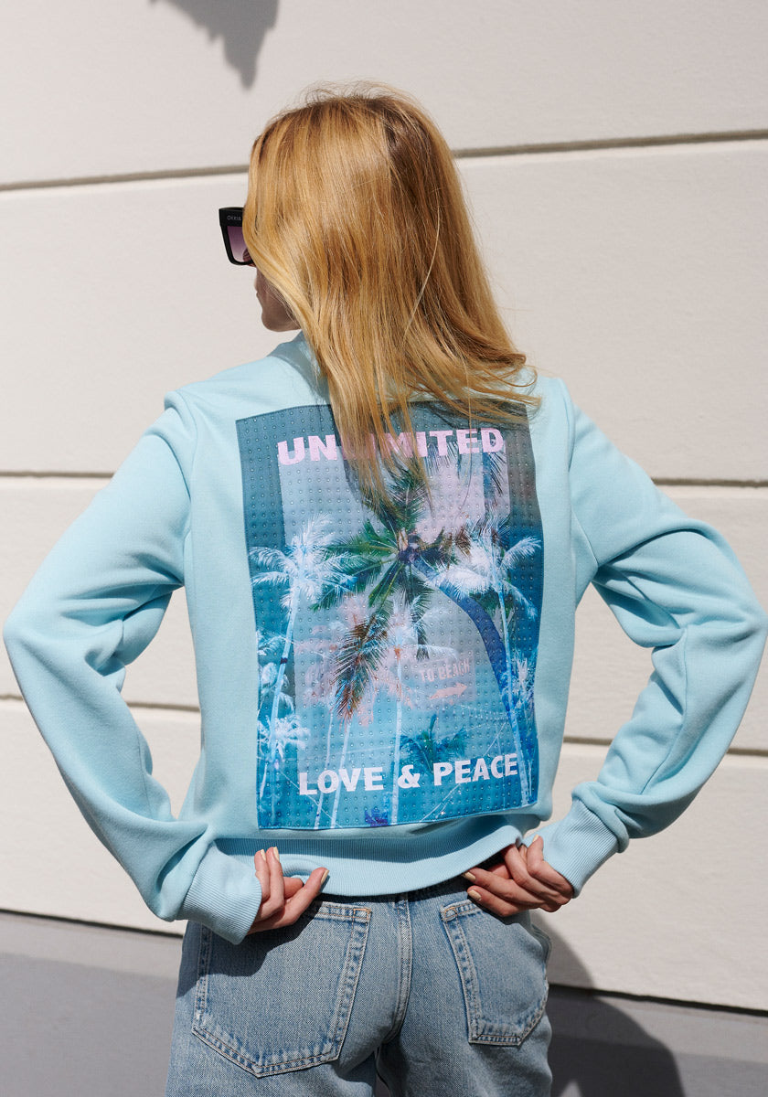 A blonde woman in sunglasses, jeans, and a light blue Princess Goes Hollywood Sweatjacke with Stehkragen stands with her back to the camera. The jacket has a colorful tropical palm tree graphic and “UNLIMITED LOVE & PEACE” on the back.