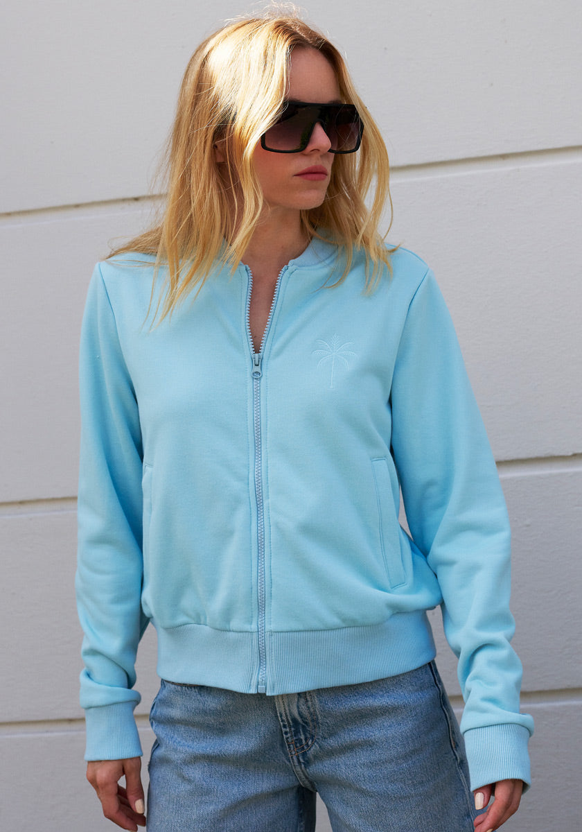 A woman with long blonde hair wears oversized black sunglasses and a light blue Princess Goes Hollywood zip-up sweatshirt with a subtle palm tree design, paired with faded blue jeans. She stands against a white textured wall, gazing to the side.