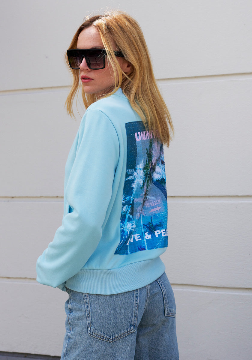 A woman with straight blonde hair wears large black sunglasses and a light blue Princess Goes Hollywood Sweatjacke featuring a palm tree graphic and “LIVE & PEACE” text on the back, paired with light blue jeans, standing against a white wall.