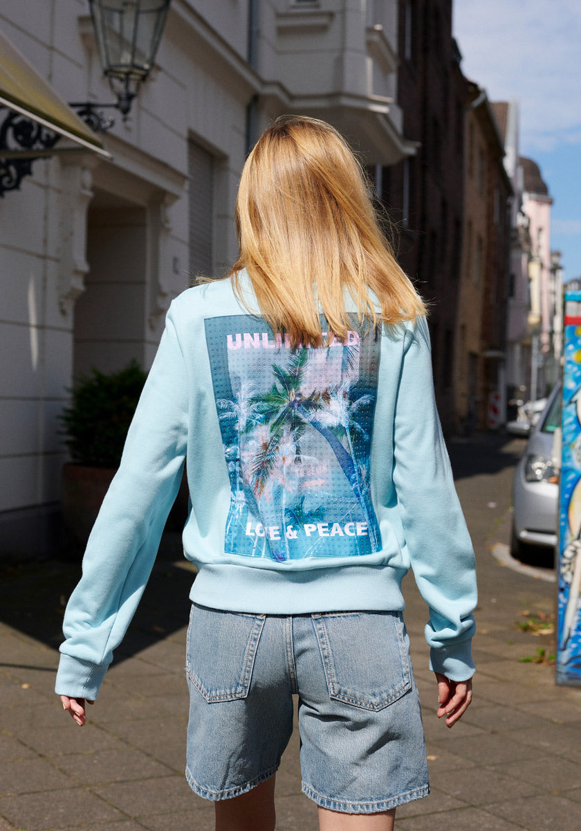 A person with straight blond hair, seen from behind, wears a light blue Princess Goes Hollywood Sweatjacke featuring palm trees and the words UNLIMITED and LOVE & PEACE, plus denim shorts, standing on a sunny, quiet city street.