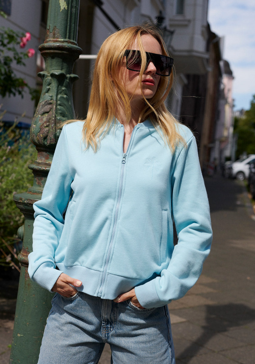 A woman with long blonde hair wears a light blue Princess Goes Hollywood Sweatjacke with Stehkragen and sunglasses, standing outdoors by a green lamppost on a city sidewalk lined with buildings and plants, hands in her jacket pockets.