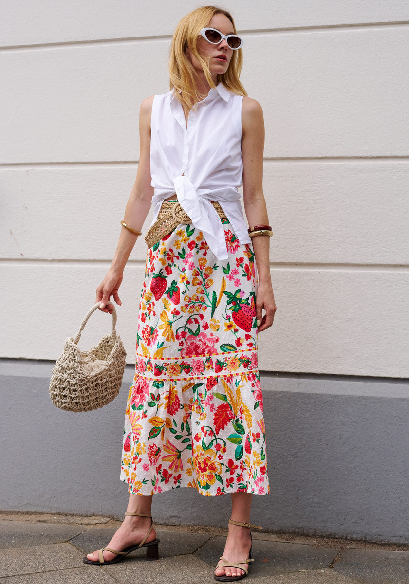 A woman in loose blonde hair wears a knotted sleeveless white shirt and a Princess Goes Hollywood Allover-Print Midirock with elastic waistband, paired with sunglasses, heeled sandals, bangles, and a woven bag against a white wall.