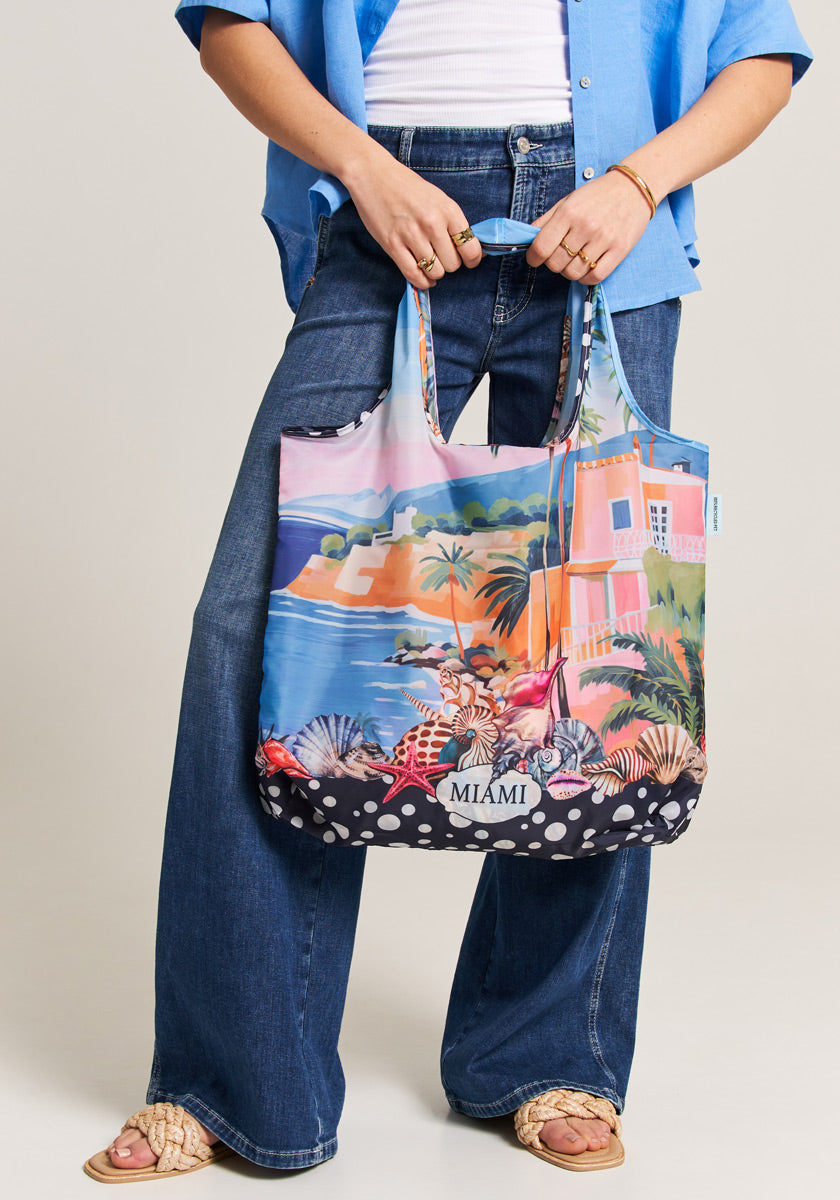 A person in a blue shirt and wide-leg jeans holds the Princess Goes Hollywood Shopper Miami, made from recycled PET with an allover Miami print—palm trees, seashells, a pink building, and “MIAMI.” They wear sandals and gold jewelry.