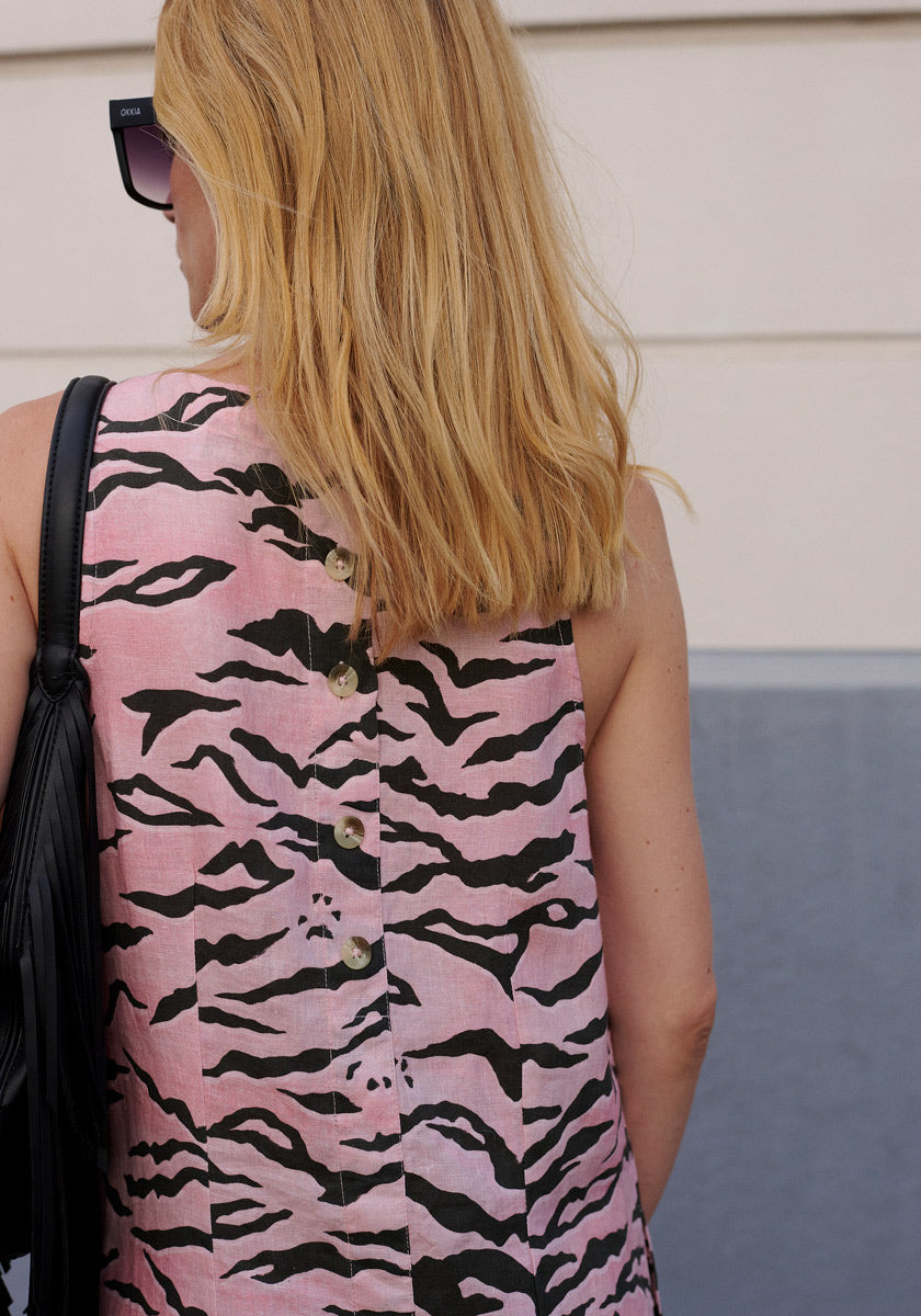 A woman with shoulder-length blonde hair is seen from behind, wearing Princess Goes Hollywood Sonnenbrille and a sleeveless pink linen tiger-print dress with back button placket. She carries a black fringed bag over her shoulder.