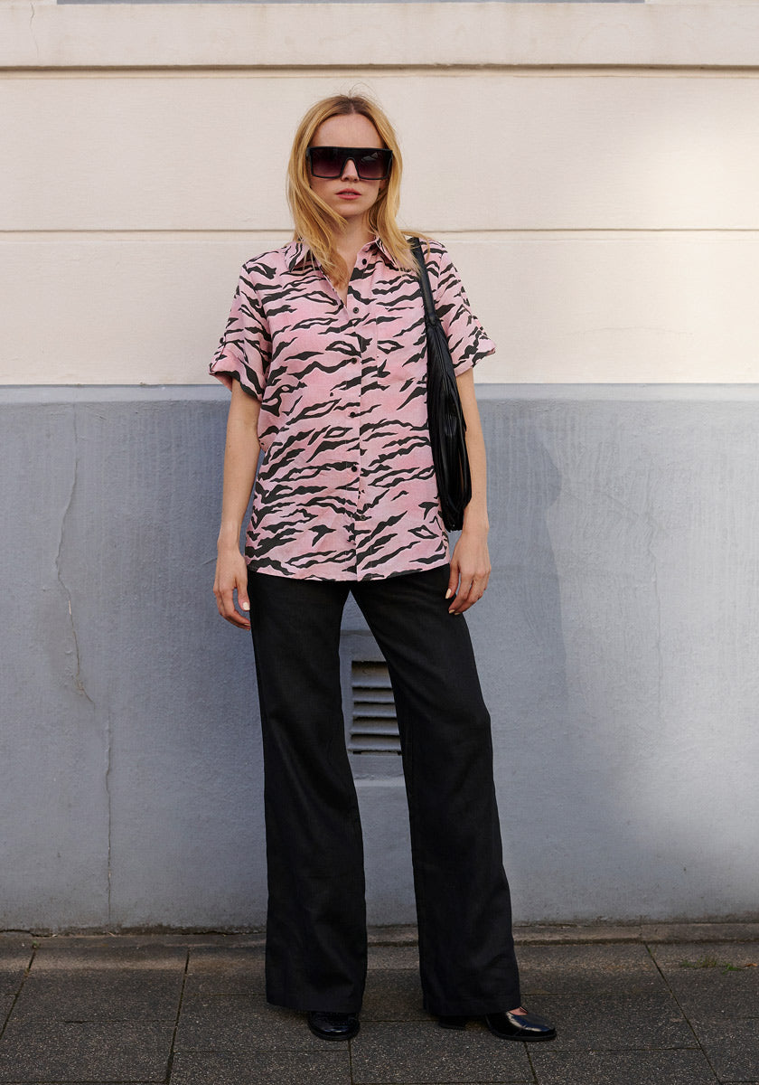 A woman stands by a two-tone wall in a loose pink and black zebra print shirt and Princess Goes Hollywood black linen pants with elastic waistband and side pockets, paired with black shoes, oversized sunglasses, and a black shoulder bag.