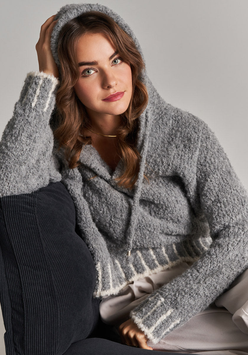 A young woman with wavy brown hair sits on a dark cushion, wearing the Grauer Strickhoodie mit Kontrastnähten by Frogbox by Princess. She gazes at the camera, left hand on her head, hood up against a neutral background.