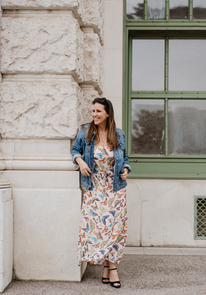 A woman stands outdoors, smiling in a blue denim jacket over a butterfly Allover-Print Midikleid with V-Ausschnitt by Princess Goes Hollywood, black sandals, sunglasses on her head, her long hair loose, and an old green-framed window behind her.