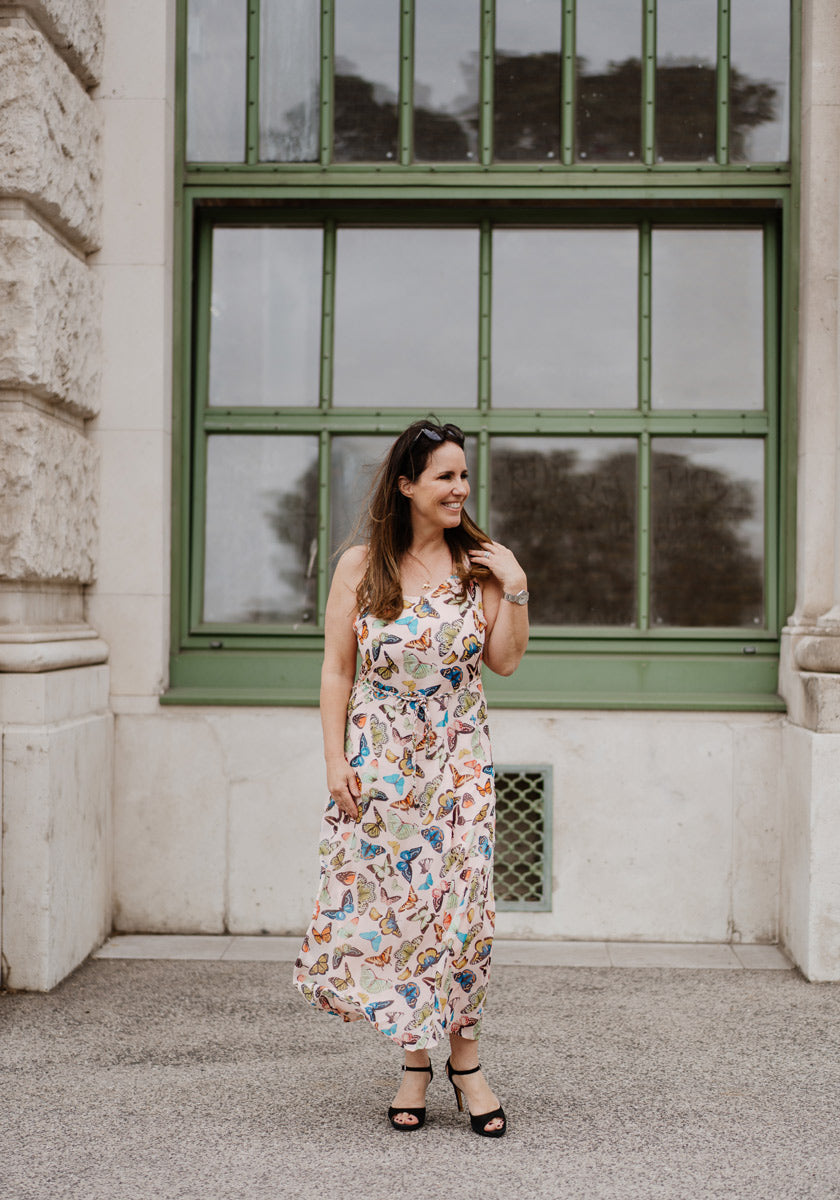 A woman in a colorful Allover-Print Midikleid by Princess Goes Hollywood stands before a large green-paned window, smiling to the side. She has long brown hair, wears sunglasses on her head, and stands on a stone walkway between light stone walls.