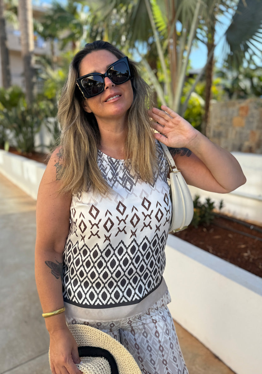 A woman with long blonde hair and sunglasses smiles outdoors, wearing a Princess Goes Hollywood sleeveless blouse with geometric print and silk blend, matching pants, holding a straw hat and white purse. Palm trees line the sunlit walkway behind her.