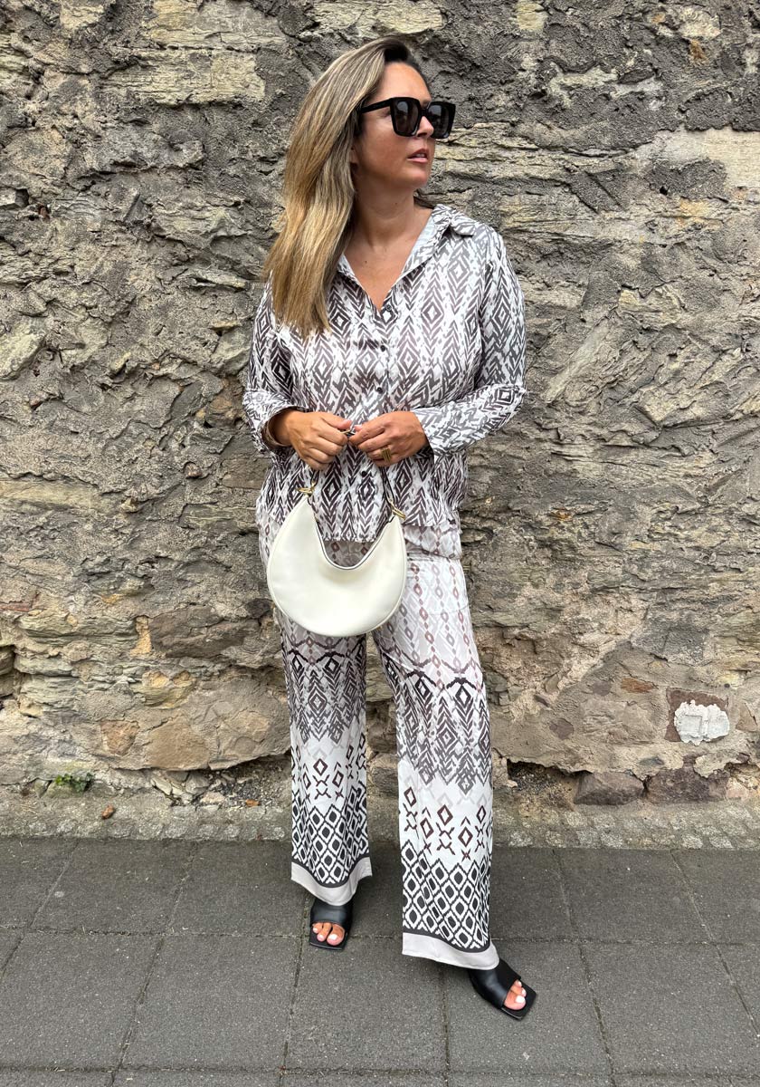 A woman with long blond hair stands by a stone wall, wearing a black-and-white geometric print Hemdbluse and pants set with silk by Princess Goes Hollywood. She carries a white crescent handbag, black sandals, and large dark sunglasses.