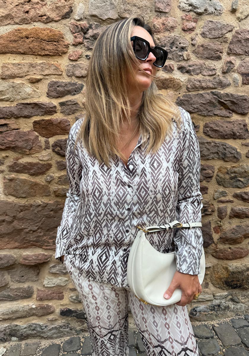 A woman with long blonde hair wears oversized black sunglasses and a Princess Goes Hollywood Hemdbluse mit Seideanteil und geometrischem Print. She holds a white handbag, standing against a textured stone wall with a confident expression.
