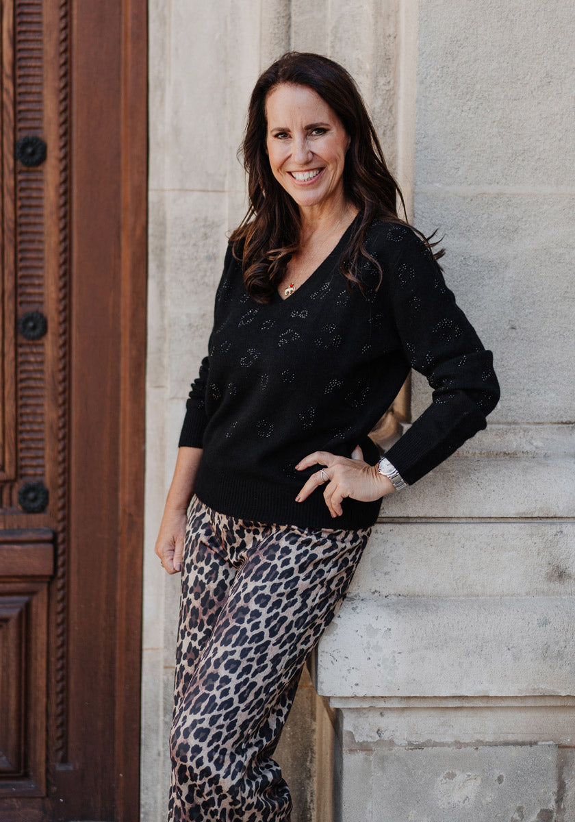A woman with long brown hair smiles by a stone wall and wooden door, wearing a Princess Goes Hollywood black Strickpullover mit Wolleanteil with subtle patterns, leopard print pants, a watch, and a necklace.