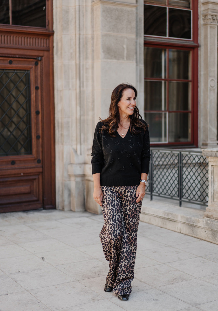 A woman with long brown hair walks outdoors, smiling. She wears a black Princess Goes Hollywood Strickpullover mit Wolleanteil and loose leopard-print pants. By a stone building with big windows and a wooden door, she looks relaxed and confident.