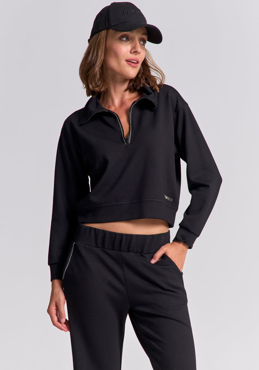 A woman with chin-length light brown hair wears a black zip-collar Sweatpullover and matching jogger pants from Princess Goes Hollywood, paired with a black baseball cap. She stands with one hand in her pocket, smiling against a neutral background.
