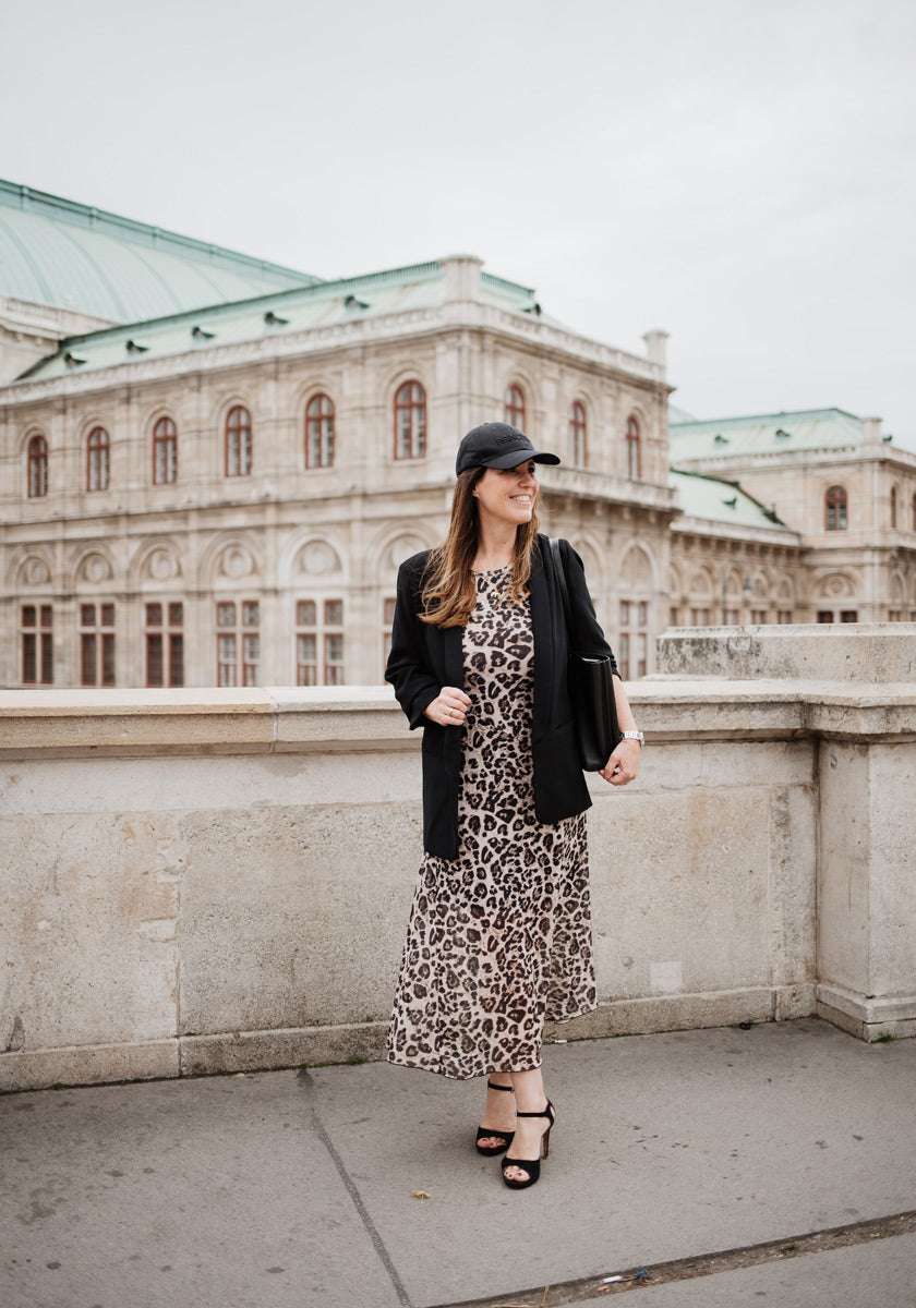 A woman stands on a stone terrace before a historic building, smiling in a black cotton cap, Princess Goes Hollywood blazer, leopard print dress, black sandals, and carrying a black bag.