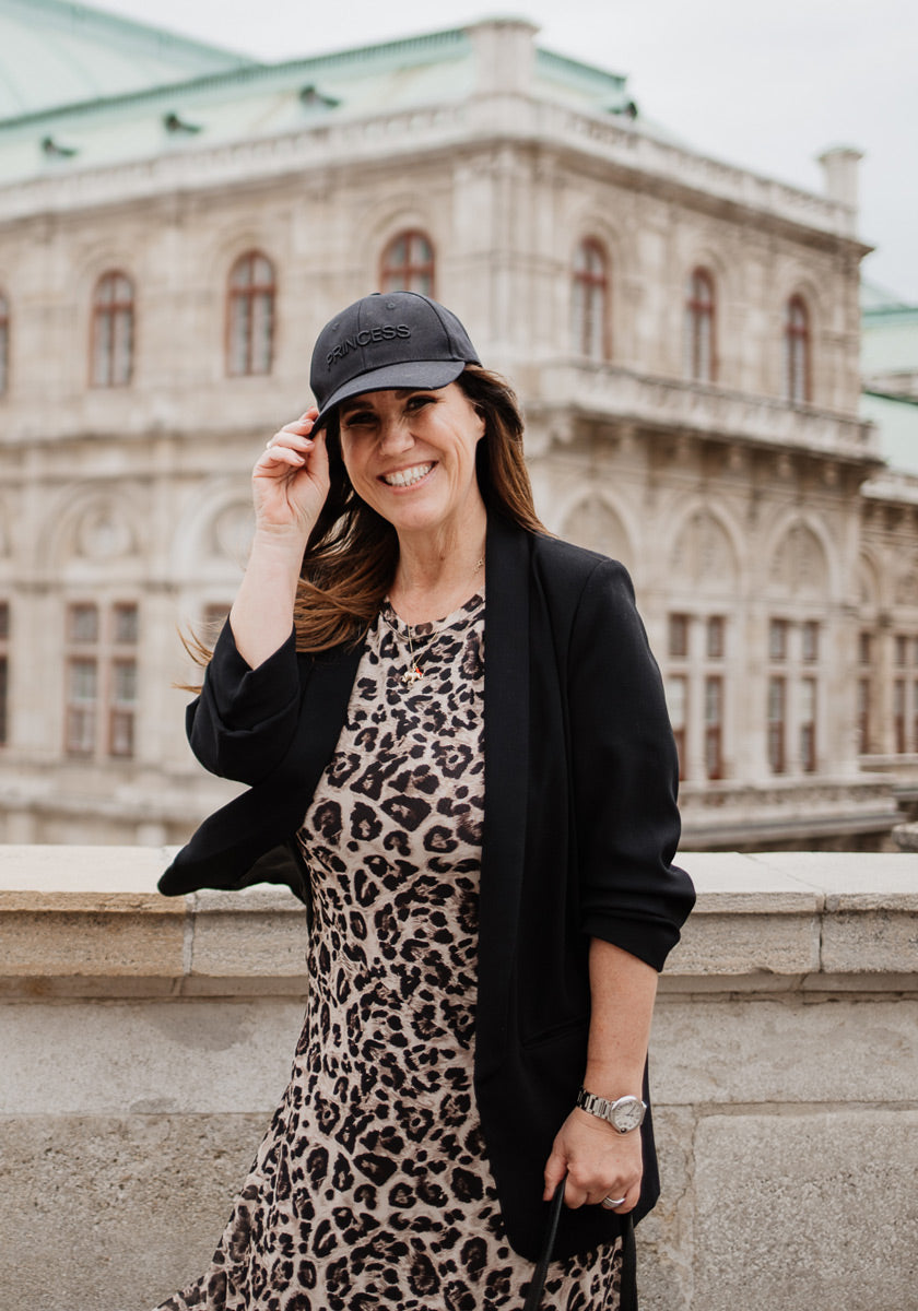 A smiling woman stands outside an ornate stone building in a black cotton cap, black blazer, leopard-print dress from Princess Goes Hollywood, and a silver watch. She holds her cap’s brim with one hand and has long dark hair.