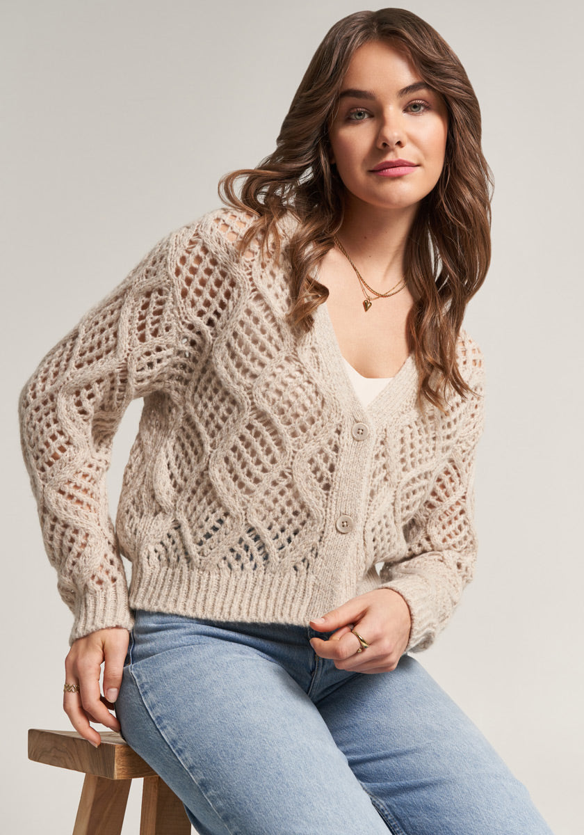 A woman with long brown hair sits on a wooden stool against a plain background, wearing a cream-colored ajour-knit cardigan by Princess Goes Hollywood over a white top and light blue jeans, accessorized with gold hoop earrings, a necklace, and a gentle smile.