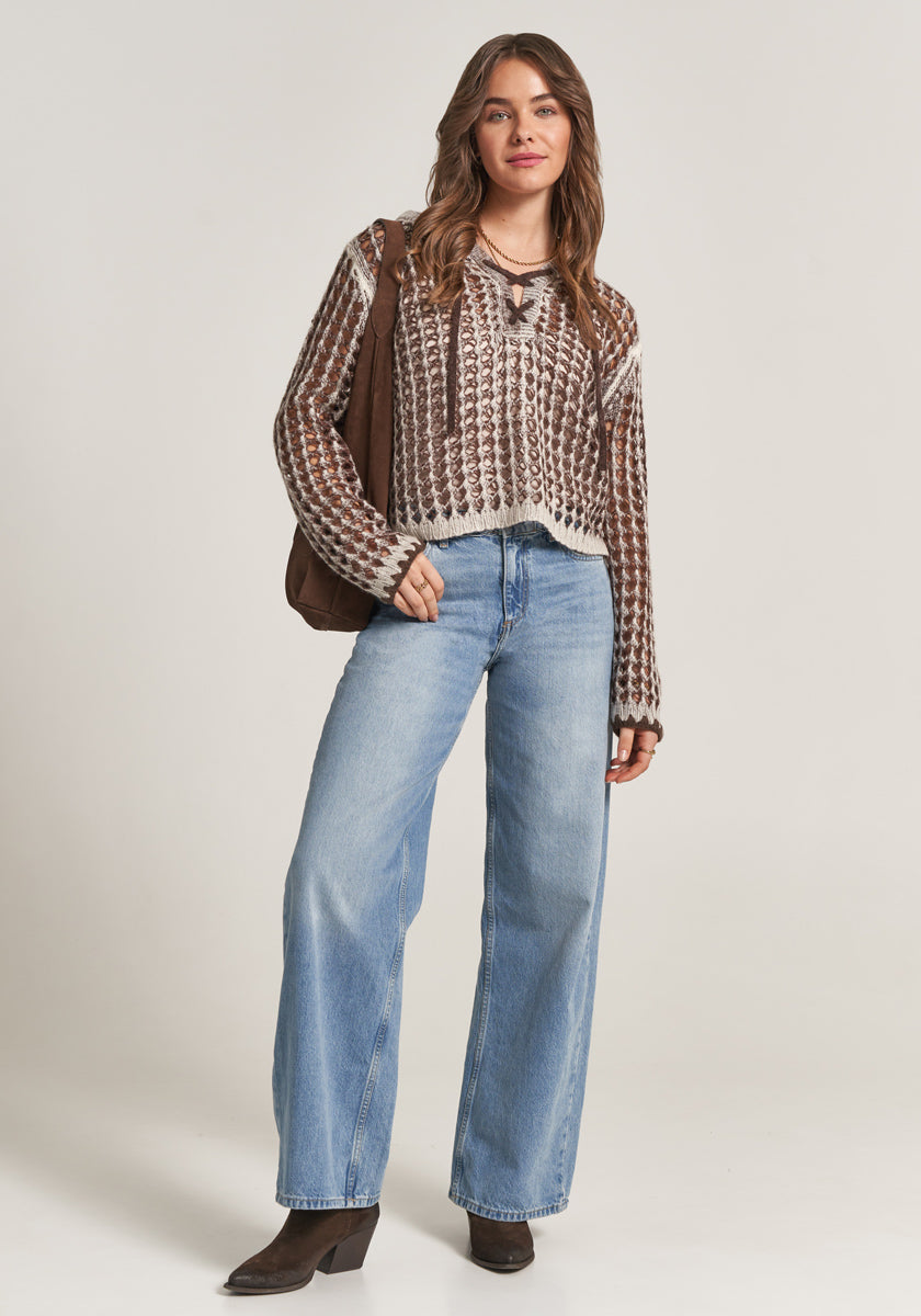 A woman with loose, wavy hair stands against a plain background, smiling softly. She wears a chunky brown and cream Ajourstrick sweater by Princess Goes Hollywood, light blue high-waisted wide-leg jeans, brown heeled boots, and carries a brown shoulder bag.