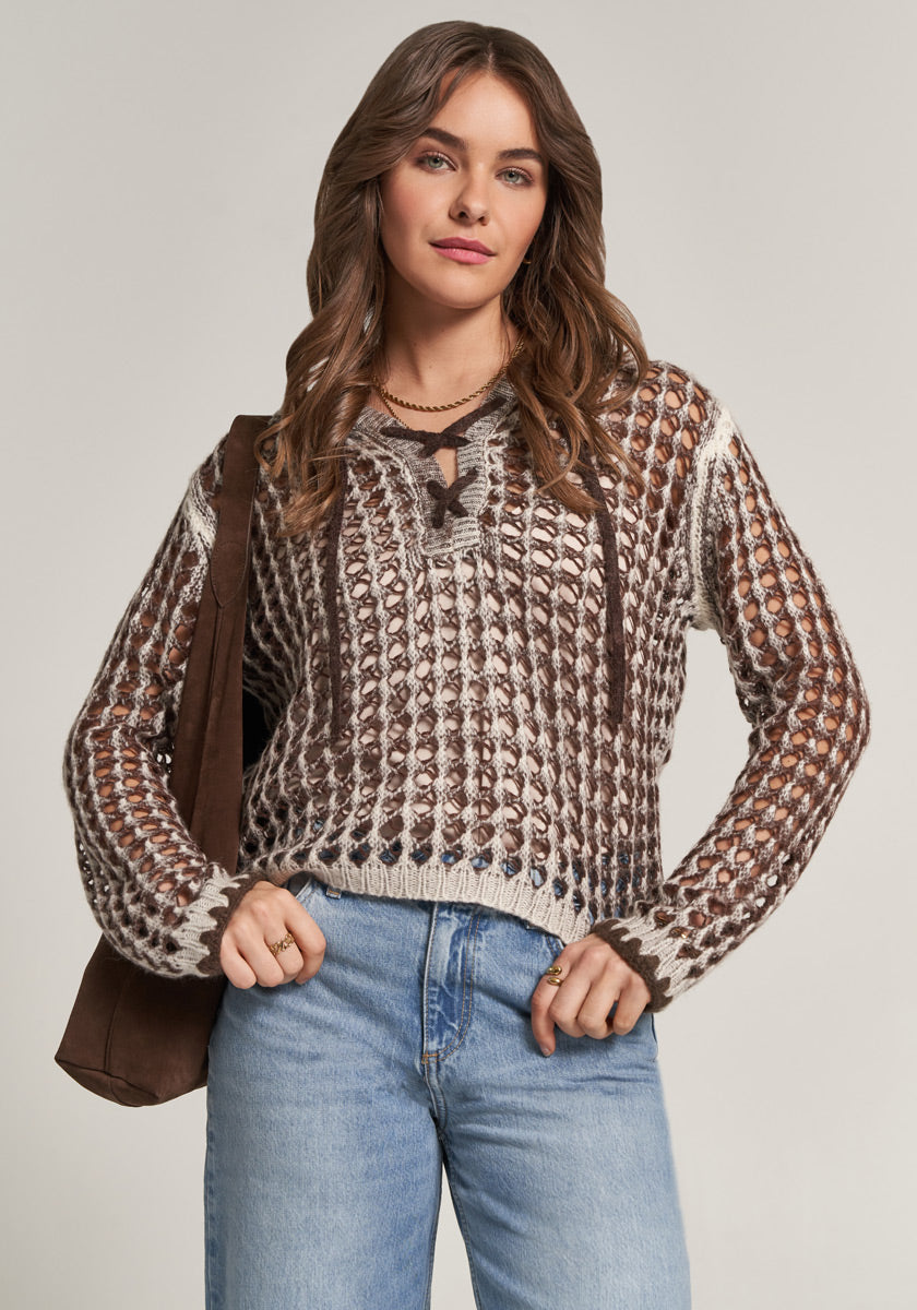 A woman with wavy brown hair wears a chunky, open-knit brown and cream sweater with a lace-up neckline from Princess Goes Hollywood, light blue jeans, and a brown shoulder bag. She stands against a plain background, smiling confidently at the camera.