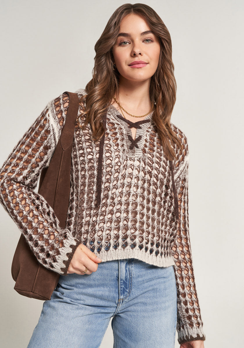 A woman with wavy brown hair wears a Princess Goes Hollywood open-knit sweater over a tank top, light blue high-waisted jeans, and carries a brown shoulder bag. She smiles slightly, standing against a plain light background.