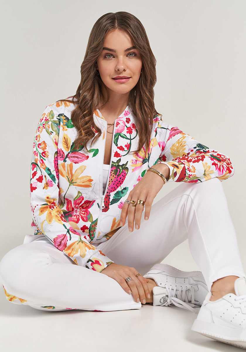 A woman with wavy brown hair sits cross-legged on the floor, smiling softly. She wears the Sweatjacke Erdbeere by Princess Goes Hollywood, paired with white pants and sneakers against a plain light background.