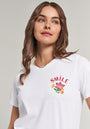 A woman with wavy brown hair wears a Princess Goes Hollywood white cotton t-shirt featuring a floral print and red “SMILE” embroidery. She smiles softly, looks upward, and pairs it with a gold chain necklace against a plain background.
