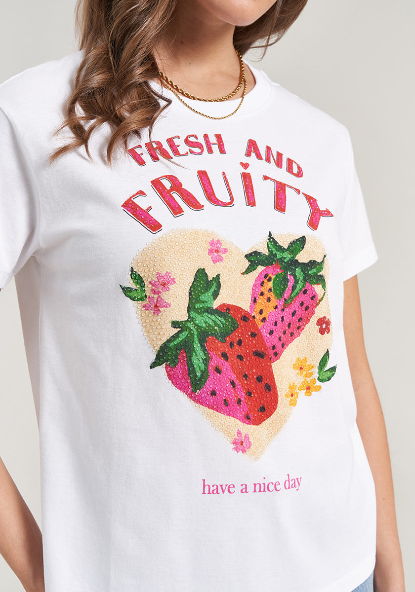 A woman wears the Princess Goes Hollywood T-Shirt Erdbeere, a white cotton tee with colorful strawberry and flower illustrations inside a pale heart, “FRESH AND FRUITY” on top, and “have a nice day” in pink and red at the bottom.