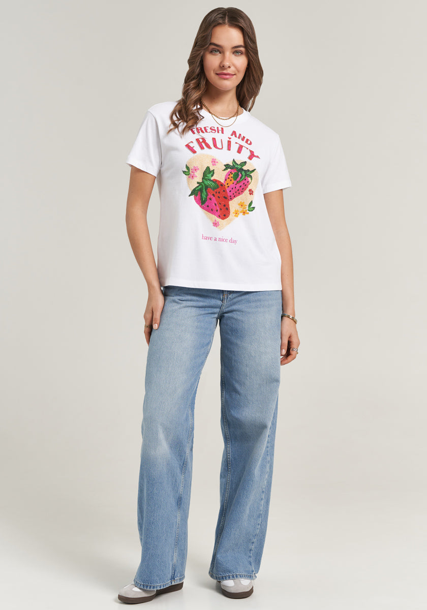 A woman with wavy brown hair smiles in a Princess Goes Hollywood white Baumwolle T-Shirt with “Fresh and Fruity” text and fruit graphics, light blue wide-leg jeans, and gray sneakers, standing against a plain light background.