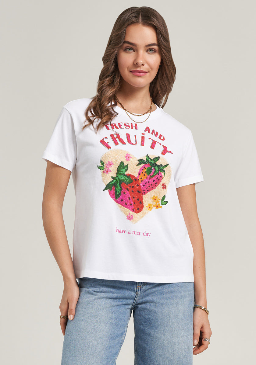 A young woman with wavy brown hair wears a white Princess Goes Hollywood Baumwolle T-Shirt featuring colorful strawberries and the phrases “Fresh and Fruity” and “have a nice day.” She pairs it with light blue jeans, posing against a plain background.