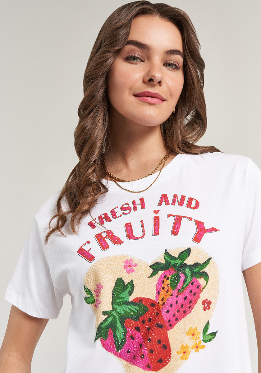 A young woman with wavy brown hair wears the Princess Goes Hollywood T-Shirt Erdbeere: a white Baumwolle tee with two large strawberries and colorful flowers in a pastel heart, topped by bold red “FRESH AND FRUITY” text. She smiles and wears a gold necklace.