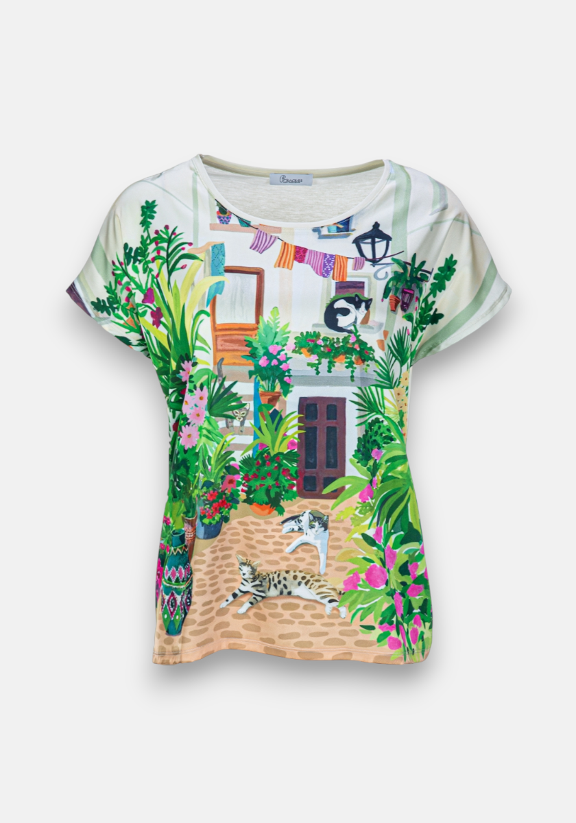 The Princess Goes Hollywood T-Shirt Garden features a vibrant Mediterranean courtyard print with colorful flowers, two lounging cats, and laundry above, creating a charming summer vibe.