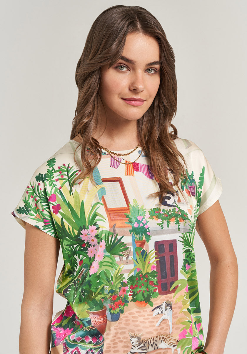 A woman with wavy brown hair and light eyes smiles in a Princess Goes Hollywood T-Shirt Garden with a lush plant, flower, red door, and two cat print, featuring a Rundhalsausschnitt. The background is pale gray.