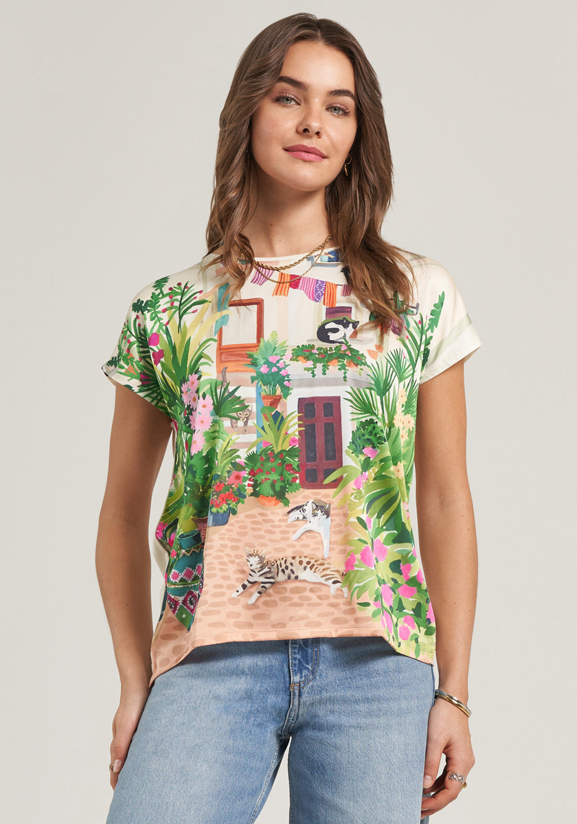 A woman with wavy brown hair wears a Princess Goes Hollywood Garten-Print T-Shirt, featuring a house, plants, and a lounging cat. She stands against a plain background, facing forward with her left thumb in her jeans pocket.