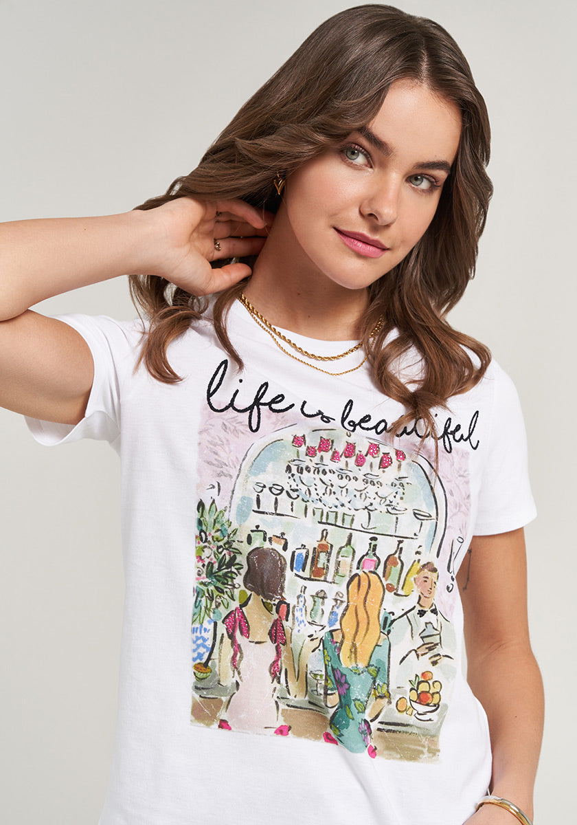 A woman with wavy brown hair wears the Princess Goes Hollywood T-Shirt Life is Beautiful, featuring a vibrant bar scene and life is beautiful script, styled with a classic Rundhalsausschnitt as she smiles gently at the camera.